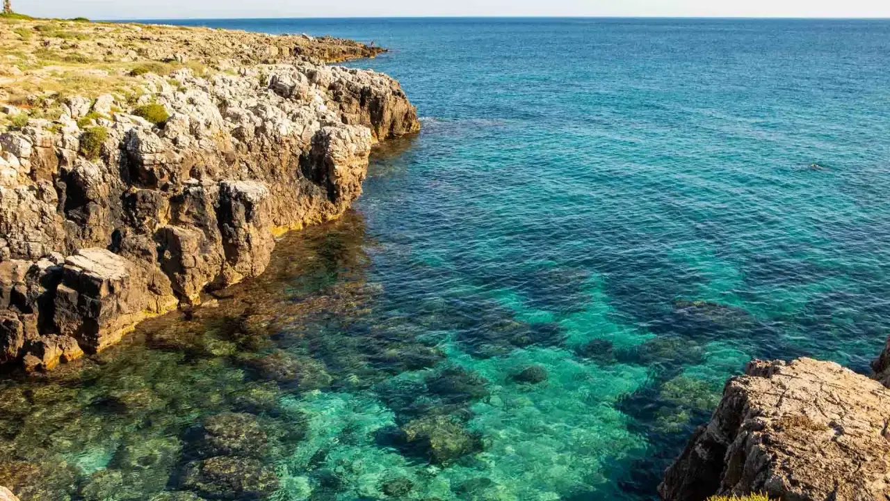 Torre solitaria su una scogliera rocciosa che domina un mare cristallino. La spiaggia sabbiosa è nascosta tra le rocce.