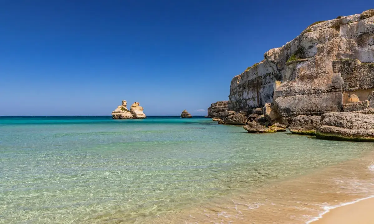 Spiaggia dorata e mare cristallino in Puglia, con faraglioni che emergono dalle acque turchesi e scogliere imponenti.