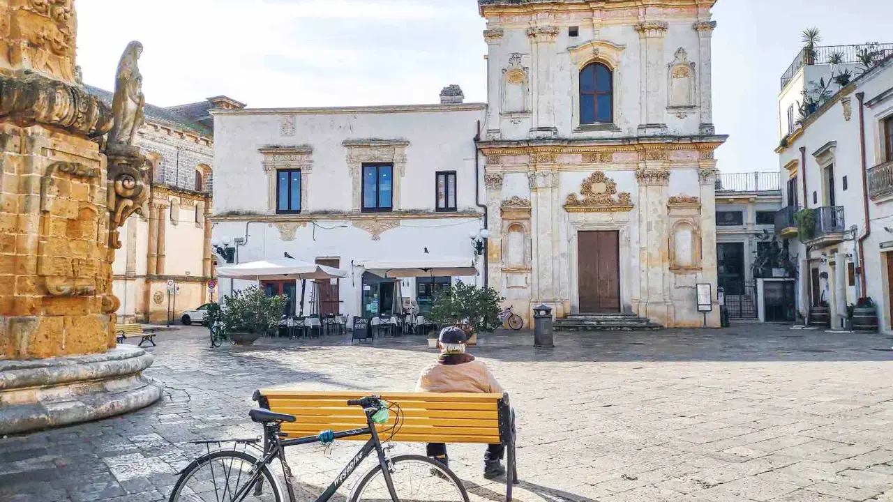 Un uomo seduto su una panchina nel **pescoluse centro storico**, con una bicicletta appoggiata. Sullo sfondo, una chiesa barocca e un caffè all'aperto.
