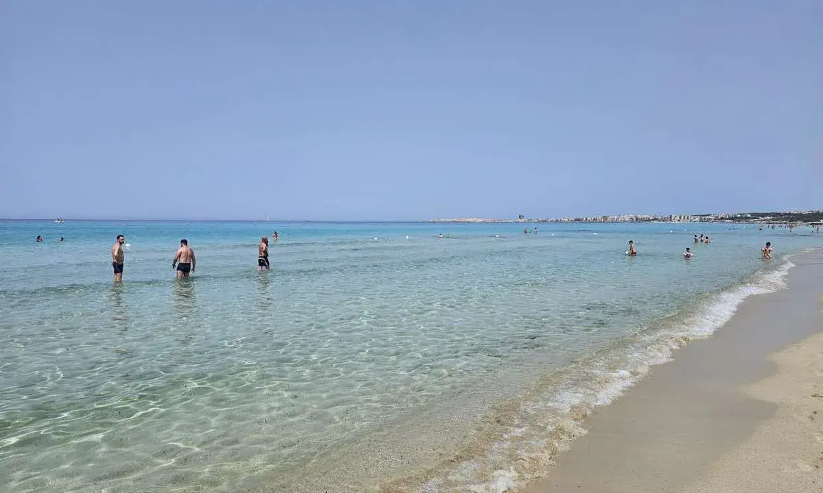 Persone in acqua cristallina vicino alla spiaggia di Gallipoli paese. Il mare calmo riflette il cielo azzurro.