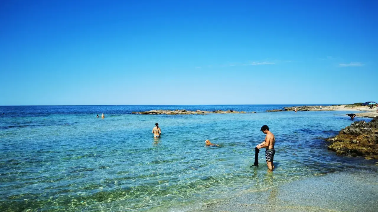 Spiaggia calette di Salve con gente che nuota e un cane che gioca nell'acqua cristallina sotto un cielo azzurro.