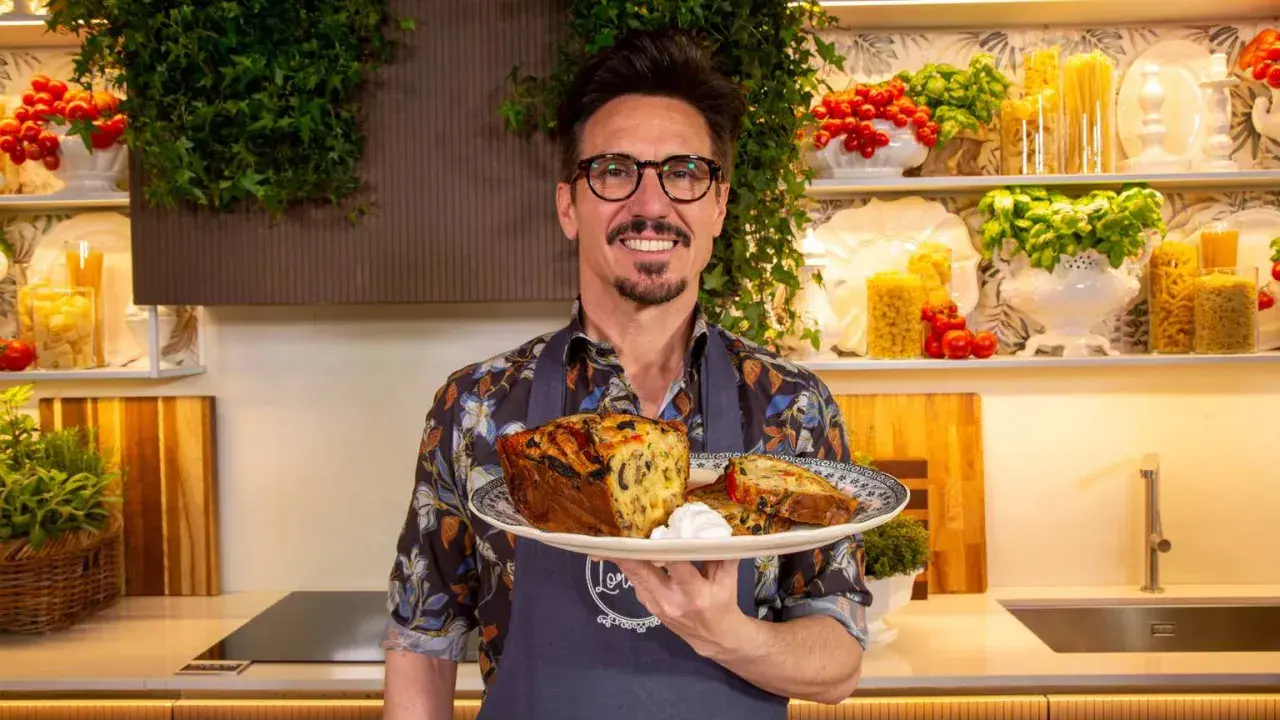 Chef sorridente con un piatto di torta salata, ispirata alle ricette leccesi, con pomodorini e basilico sullo sfondo.