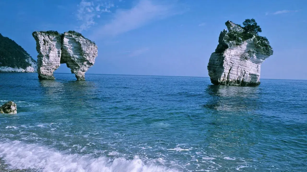 La **Baia delle Zagare** con le sue iconiche faraglioni bianchi che emergono dal mare blu intenso, sotto un cielo limpido.