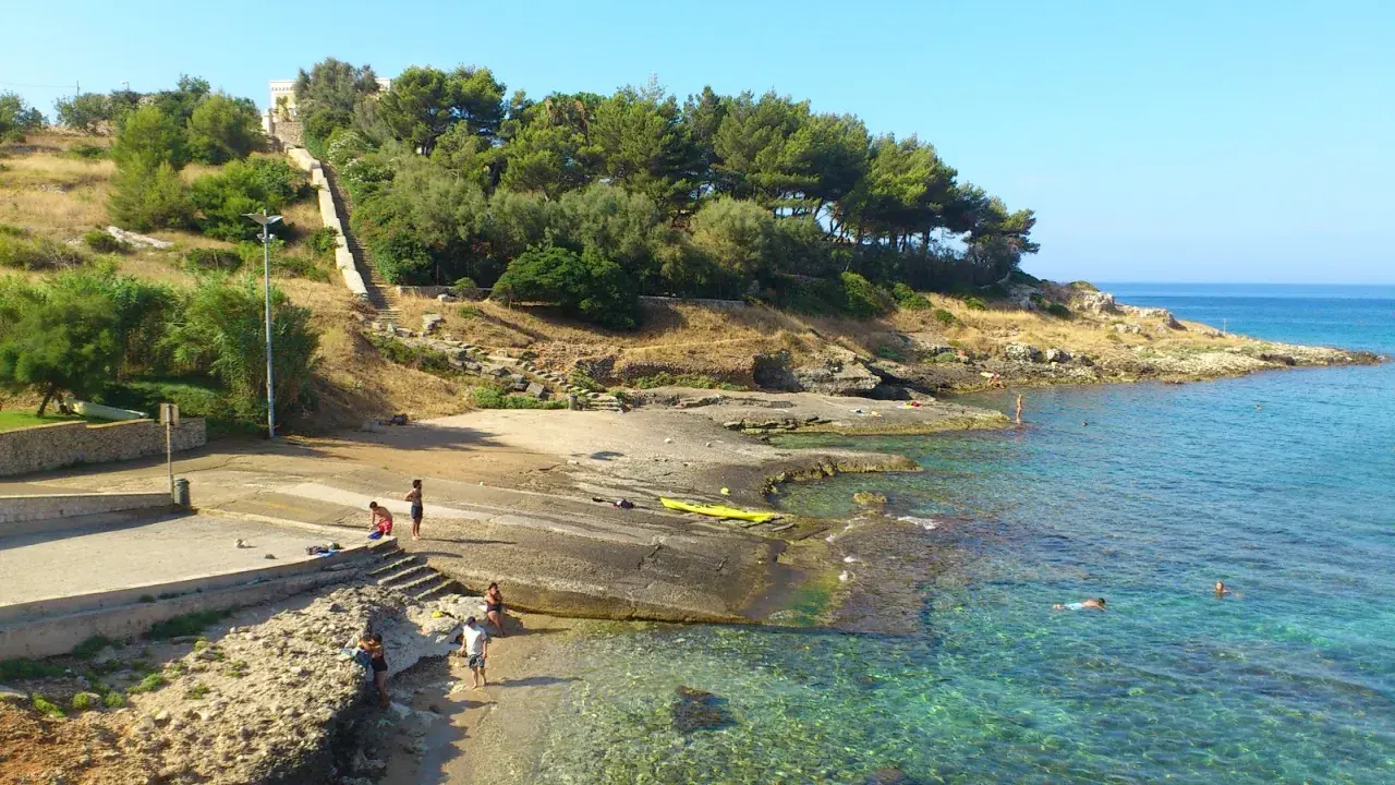 Scogliera e mare cristallino a San Gregorio, Puglia. Persone si godono il sole e l'acqua, con una scalinata che sale verso la vegetazione.