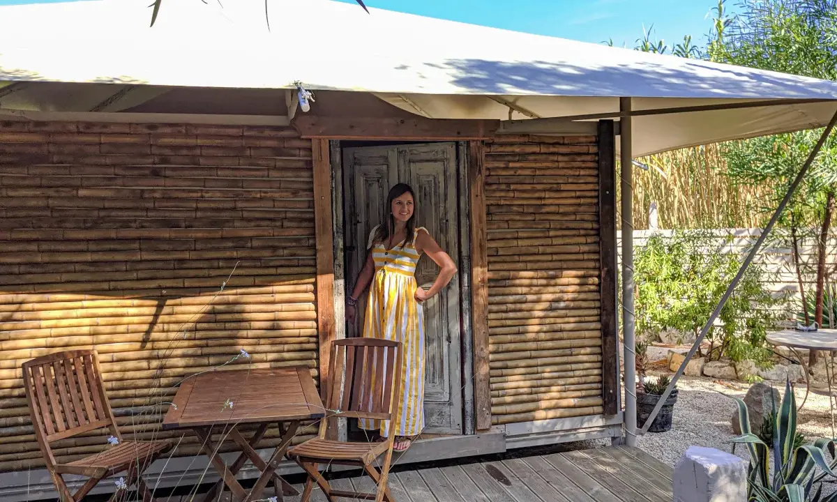 Donna sorridente in un abito a righe gialle davanti a una tenda di bambù, che incarna il glamping significato: lusso e natura.