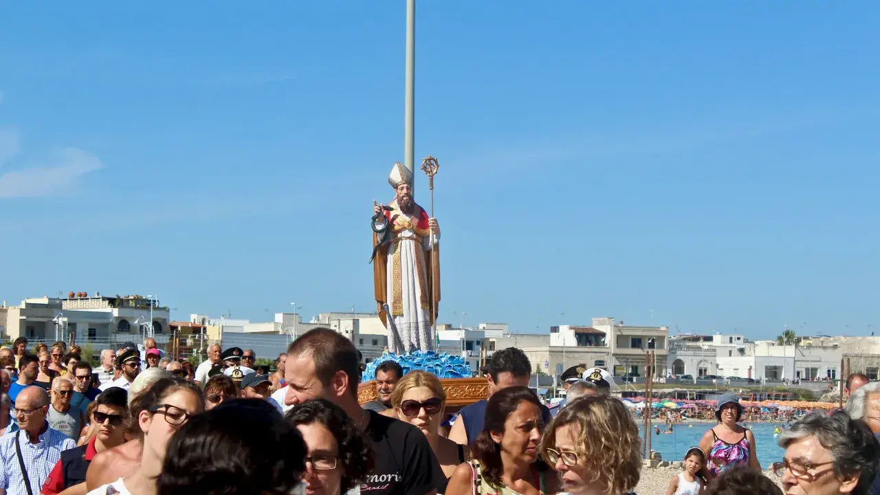 Festa San Foca: folla partecipa alla processione con la statua del santo su un carro, sotto un cielo azzurro.