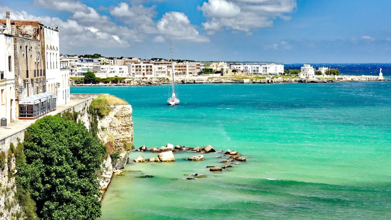 Vista mozzafiato del paese di Otranto con le sue case bianche affacciate sul mare turchese e un veliero solitario.