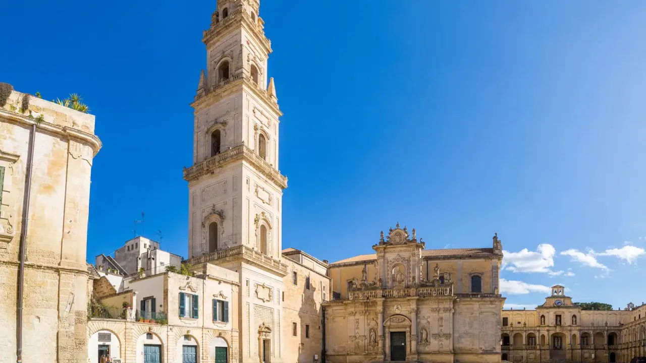 La piazza Carmelo Bene a Lecce, con la maestosa Basilica di Santa Croce e il campanile che svetta sotto un cielo azzurro.