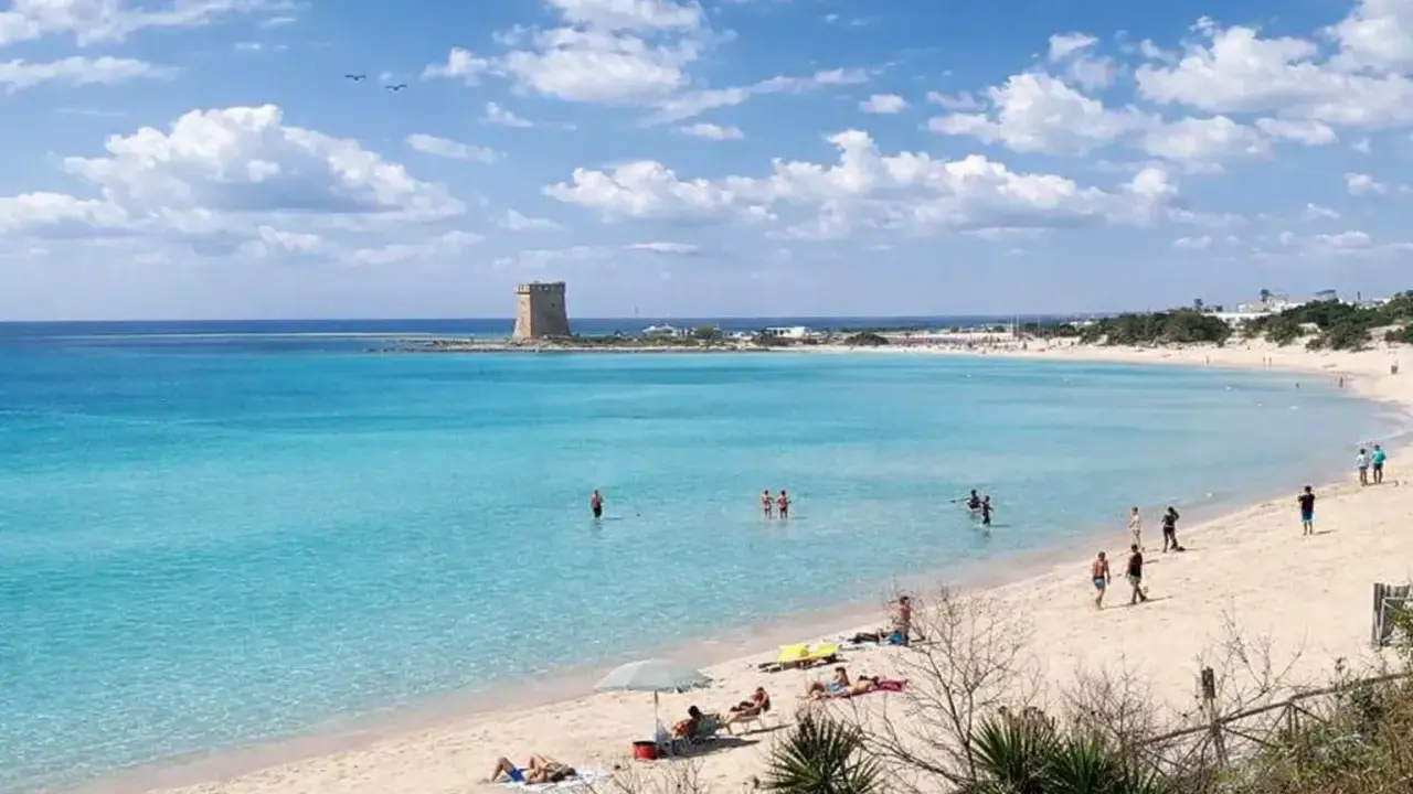 Spiaggia Scala di Furno con torre antica e acque turchesi. Persone si godono il sole e il mare cristallino.