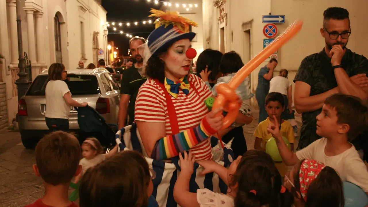 Clown con palloncino arancione anima la sagra di paese, circondata da bambini curiosi e adulti festanti.