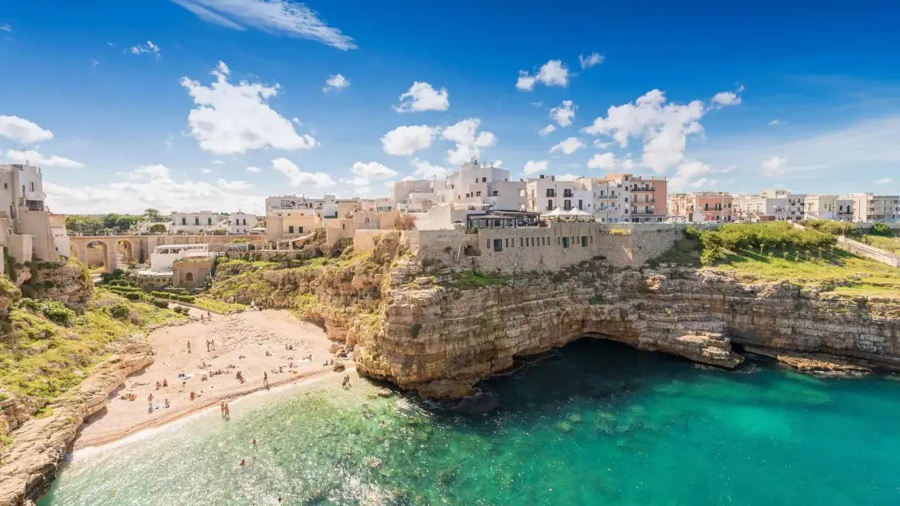 Un borgo bianco su scogliere a picco sul mare turchese, una spiaggia affollata e un invito a scoprire la Puglia da visitare.