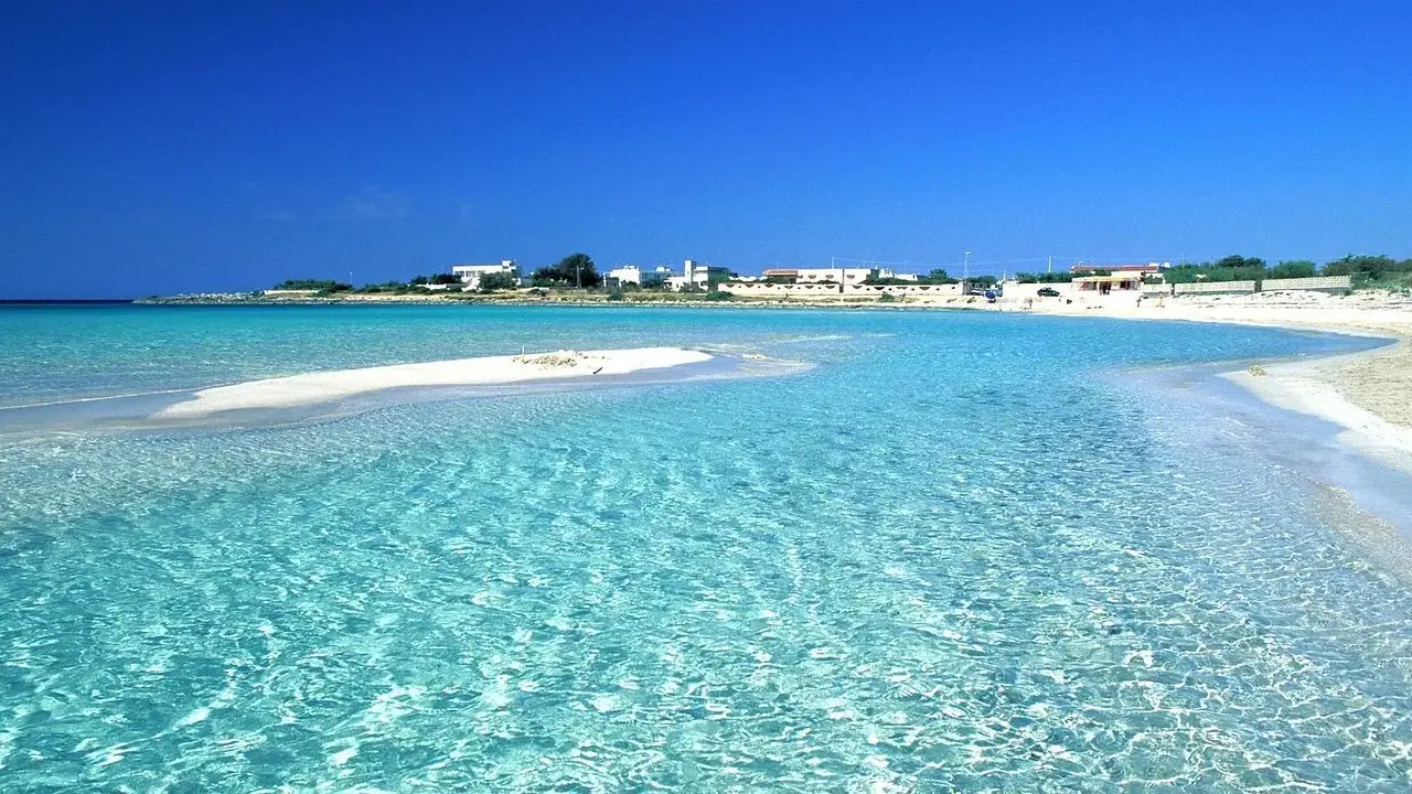 Acque cristalline lambiscono spiagge bianche, con un villaggio sullo sfondo sotto un cielo azzurro intenso.
