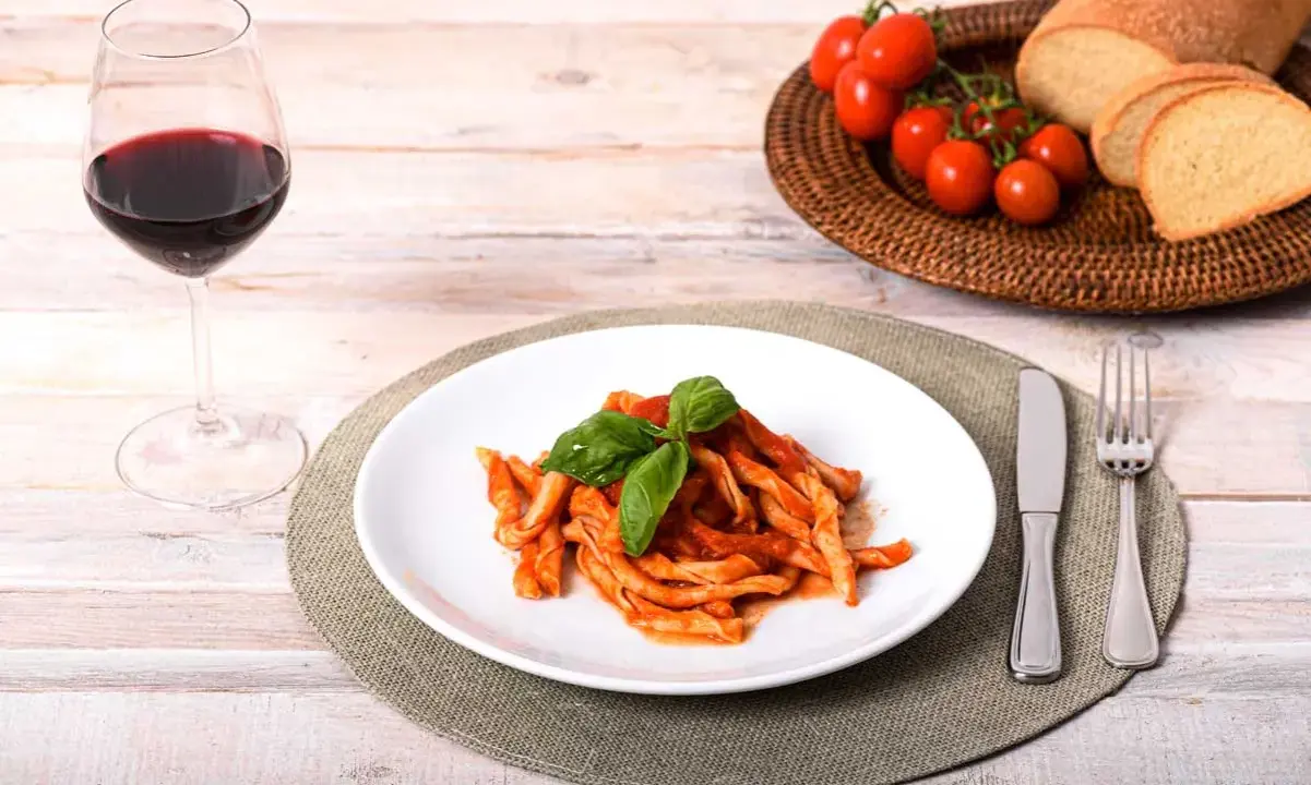 Piatto di sagne ncannulate al sugo, guarnite con basilico, accompagnate da un bicchiere di vino rosso, pane e pomodorini.