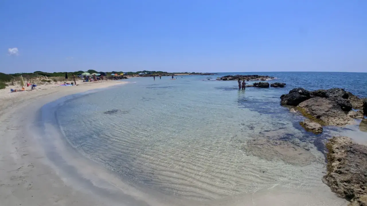 Spiaggia di sabbia bianca e mare cristallino a Marina di Salve, con scogli e persone che si godono il sole.