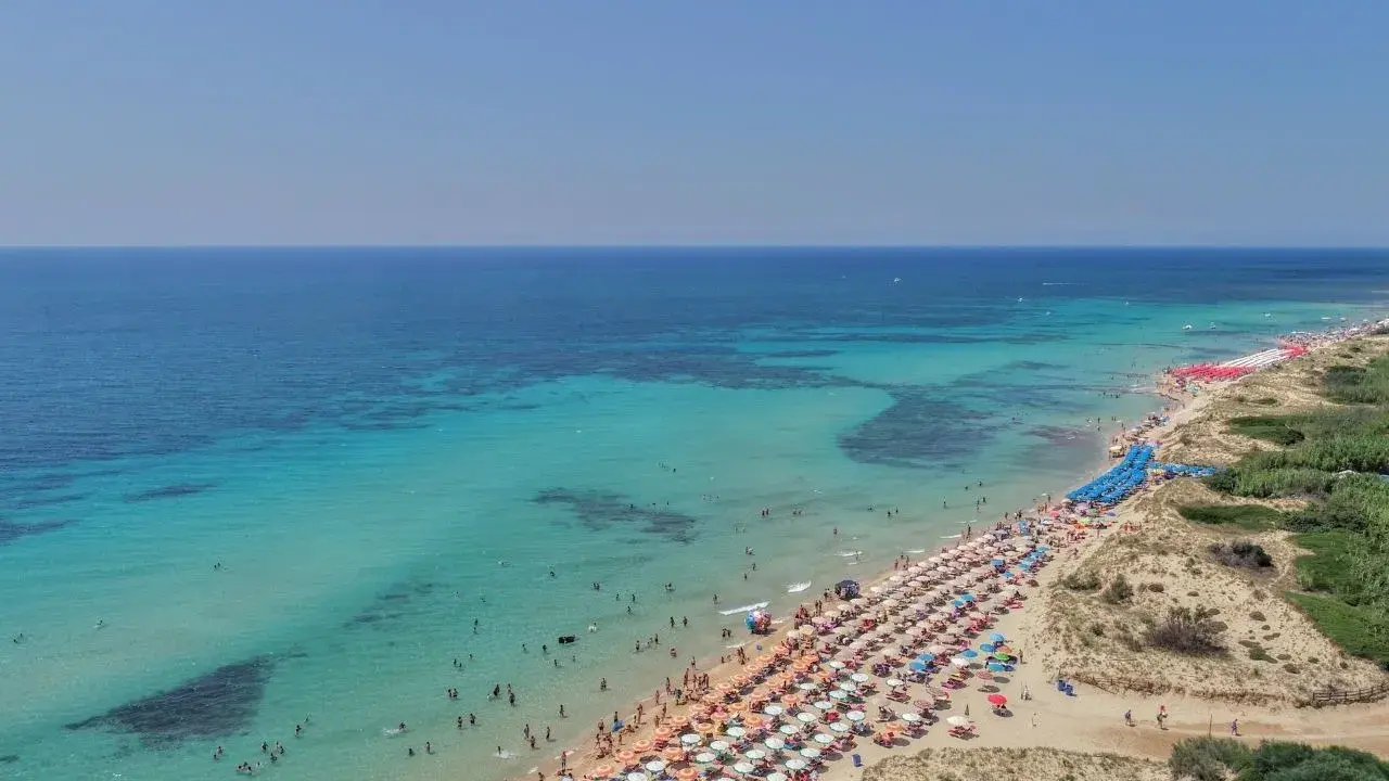 Un tratto di costa con acque cristalline e sabbia dorata, tra le spiaggie più belle del Salento, affollata di ombrelloni e bagnanti.