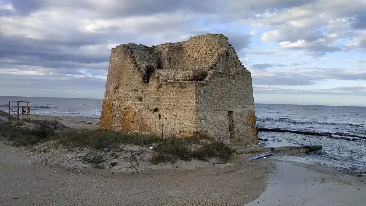 Ruined stone tower on a sandy beach with the sea and cloudy sky in the background.