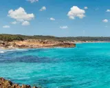 Acque cristalline e scogliere rocciose a Marina di Lecce, con una spiaggia sabbiosa e vegetazione mediterranea sullo sfondo sotto un cielo azzurro con nuvole bianche.