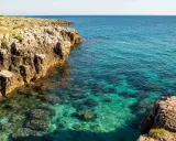 Torre solitaria su una scogliera rocciosa che domina un mare cristallino. La spiaggia sabbiosa è nascosta tra le rocce.
