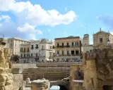 Antiche rovine romane si affacciano su palazzi barocchi nel centro storico di Lecce, sotto un cielo azzurro con nuvole bianche.