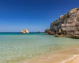 Spiaggia dorata e mare cristallino in Puglia, con faraglioni che emergono dalle acque turchesi e scogliere imponenti.