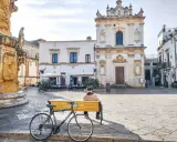 Un uomo seduto su una panchina nel **pescoluse centro storico**, con una bicicletta appoggiata. Sullo sfondo, una chiesa barocca e un caffè all'aperto.