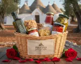 Cesto con prodotti tipici pugliesi: olio, pasta, sughi, olive e peperoncini, con rose rosse e trulli sullo sfondo.