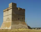 Torre costiera a Sant Isidoro, Salento. Antica sentinella di pietra contro il cielo azzurro, con il mare e il borgo sullo sfondo.