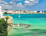Vista mozzafiato del paese di Otranto con le sue case bianche affacciate sul mare turchese e un veliero solitario.