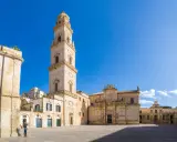 La piazza Carmelo Bene a Lecce, con la maestosa Basilica di Santa Croce e il campanile che svetta sotto un cielo azzurro.