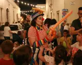 Clown con palloncino arancione anima la sagra di paese, circondata da bambini curiosi e adulti festanti.