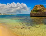 L'isola della fanciulla, una torre rotonda in pietra nel mare cristallino, con una spiaggia dorata e case in lontananza sotto un cielo azzurro con nuvole bianche.