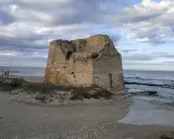 Ruined stone tower on a sandy beach with the sea and cloudy sky in the background.