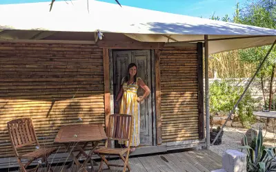 Donna sorridente in un abito a righe gialle davanti a una tenda di bambù, che incarna il glamping significato: lusso e natura.