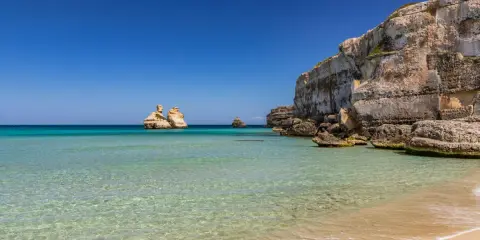 Spiaggia dorata e mare cristallino in Puglia, con faraglioni che emergono dalle acque turchesi e scogliere imponenti.