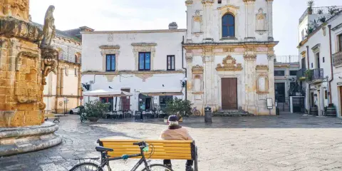 Un uomo seduto su una panchina nel **pescoluse centro storico**, con una bicicletta appoggiata. Sullo sfondo, una chiesa barocca e un caffè all'aperto.