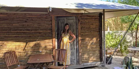 Donna sorridente in un abito a righe gialle davanti a una tenda di bambù, che incarna il glamping significato: lusso e natura.