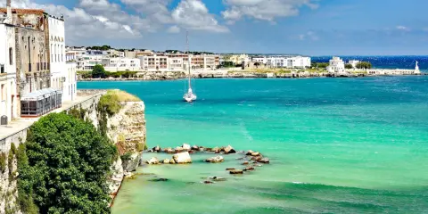 Vista mozzafiato del paese di Otranto con le sue case bianche affacciate sul mare turchese e un veliero solitario.