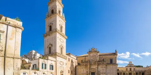 La piazza Carmelo Bene a Lecce, con la maestosa Basilica di Santa Croce e il campanile che svetta sotto un cielo azzurro.