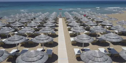 Filettato di ombrelloni di paglia e lettini sulla sabbia dorata, con il mare azzurro sullo sfondo. Un paradiso a Presicce Lido Marini.