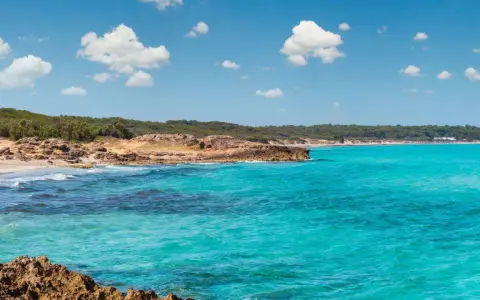 Acque cristalline e scogliere rocciose a Marina di Lecce, con una spiaggia sabbiosa e vegetazione mediterranea sullo sfondo sotto un cielo azzurro con nuvole bianche.