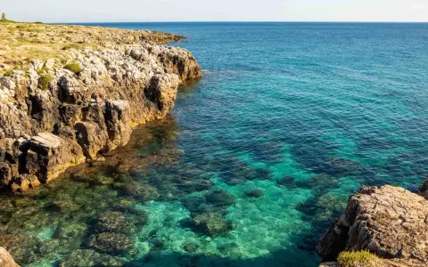 Torre solitaria su una scogliera rocciosa che domina un mare cristallino. La spiaggia sabbiosa è nascosta tra le rocce.