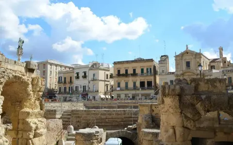 Antiche rovine romane si affacciano su palazzi barocchi nel centro storico di Lecce, sotto un cielo azzurro con nuvole bianche.