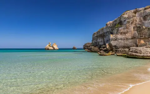 Spiaggia dorata e mare cristallino in Puglia, con faraglioni che emergono dalle acque turchesi e scogliere imponenti.