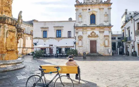 Un uomo seduto su una panchina nel **pescoluse centro storico**, con una bicicletta appoggiata. Sullo sfondo, una chiesa barocca e un caffè all'aperto.