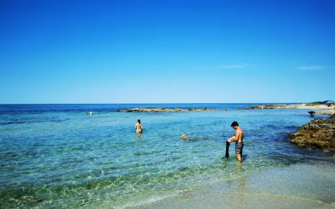 Spiaggia calette di Salve con gente che nuota e un cane che gioca nell'acqua cristallina sotto un cielo azzurro.
