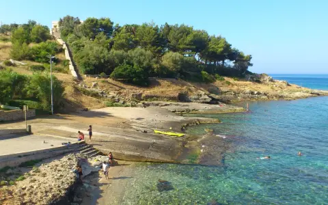 Scogliera e mare cristallino a San Gregorio, Puglia. Persone si godono il sole e l'acqua, con una scalinata che sale verso la vegetazione.