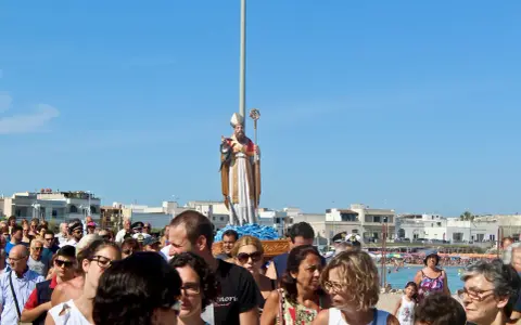 Festa San Foca: folla partecipa alla processione con la statua del santo su un carro, sotto un cielo azzurro.