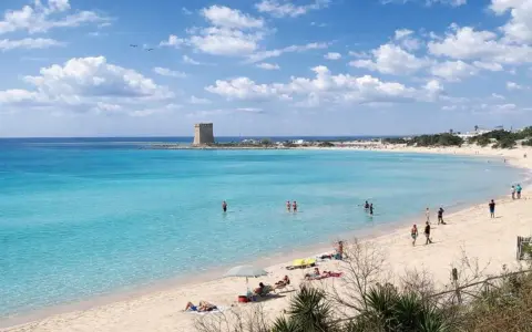 Spiaggia Scala di Furno con torre antica e acque turchesi. Persone si godono il sole e il mare cristallino.