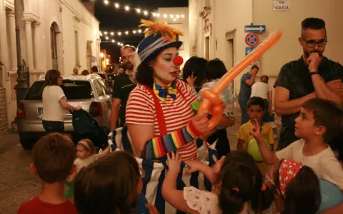 Clown con palloncino arancione anima la sagra di paese, circondata da bambini curiosi e adulti festanti.