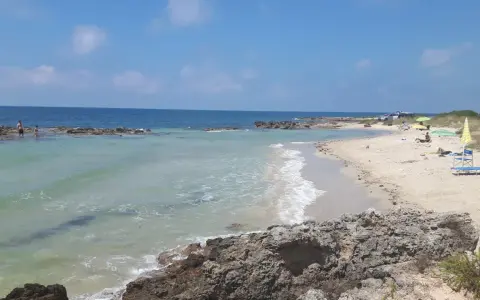 Spiaggia con scogli e mare cristallino, tipica delle calette puglia. Persone in acqua e sulla sabbia, ombrelloni e lettini.